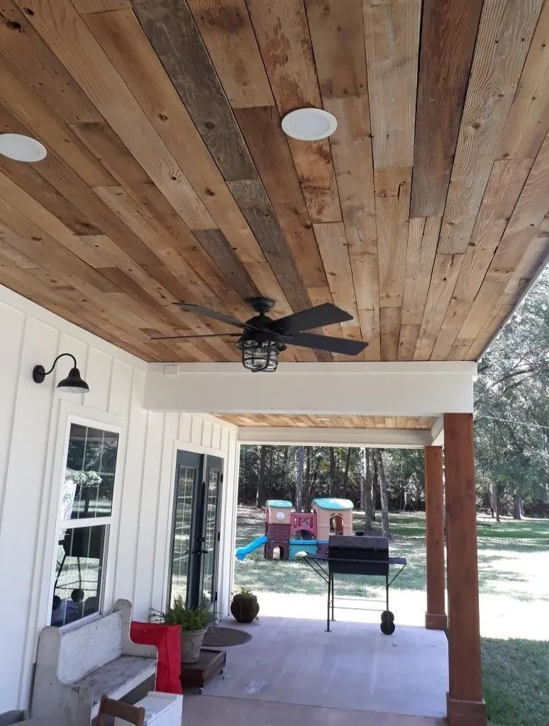 Reclaimed wood plank porch ceiling with a black ceiling fan and recessed light above a white board and batten wall