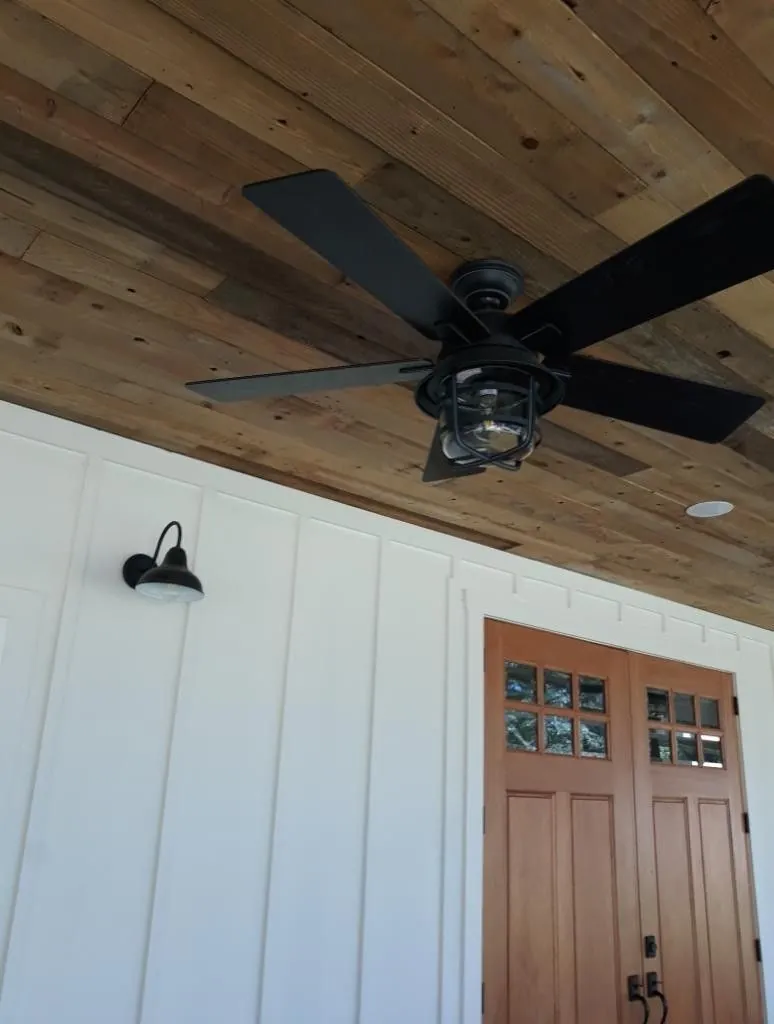 Close view of a black ceiling fan under a rustic wood plank ceiling beside a barn style wall light and double wood doors